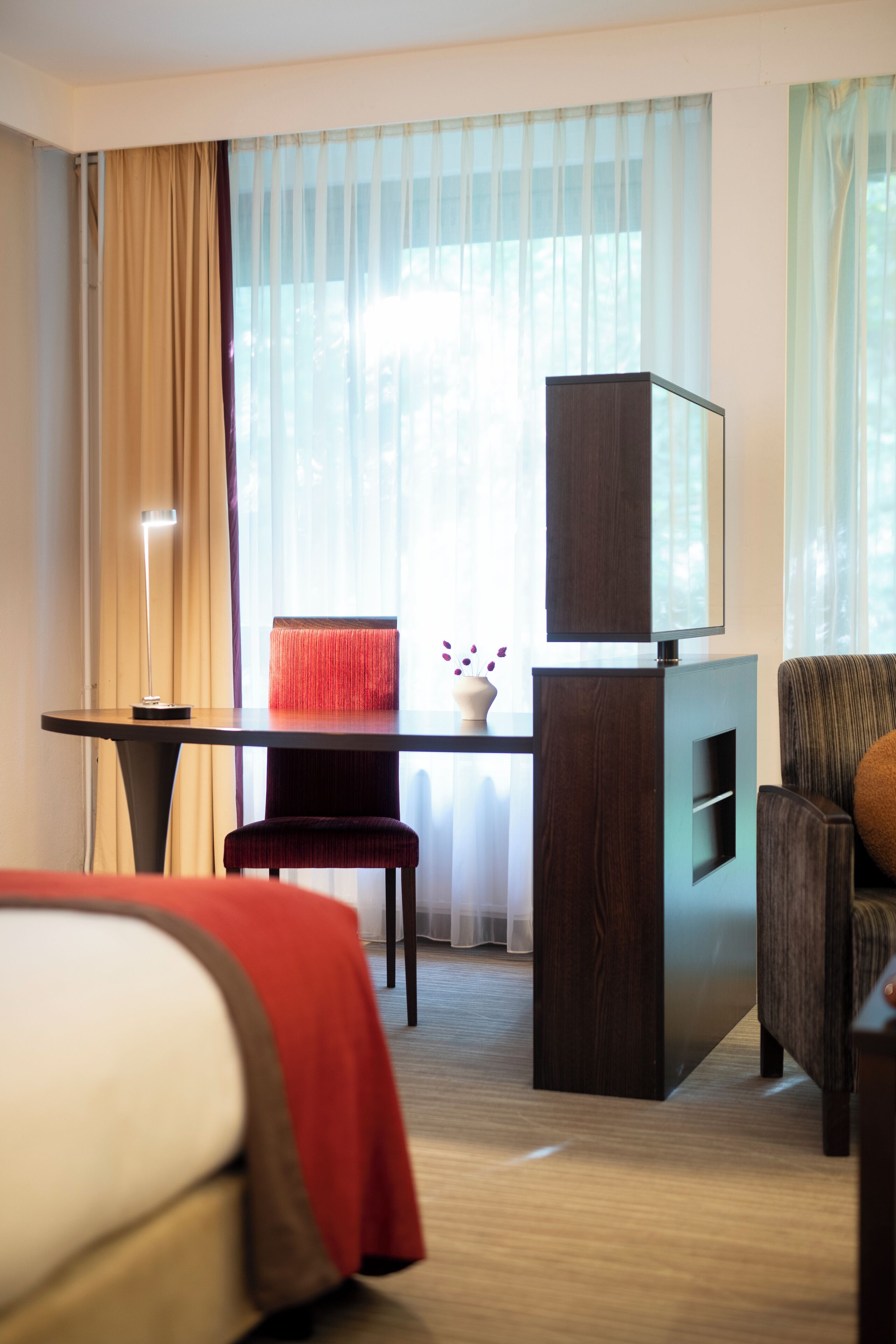 A hotel room with a bed, a desk with a television, a chair, and a window with curtains.