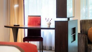 Quarto de hotel com cama, escrivaninha com televisão, cadeira e janela com cortinas.