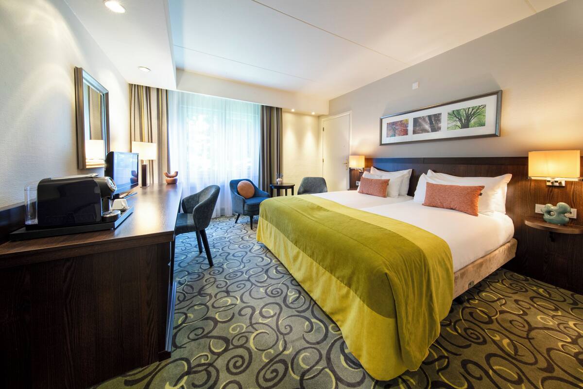 Habitación de hotel con una cama grande, un escritorio con ordenador, una silla y una mesita de noche con lámpara.