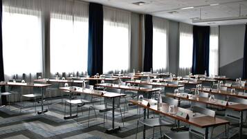 A conference room with rows of tables and chairs, large windows with curtains, and a carpeted floor.