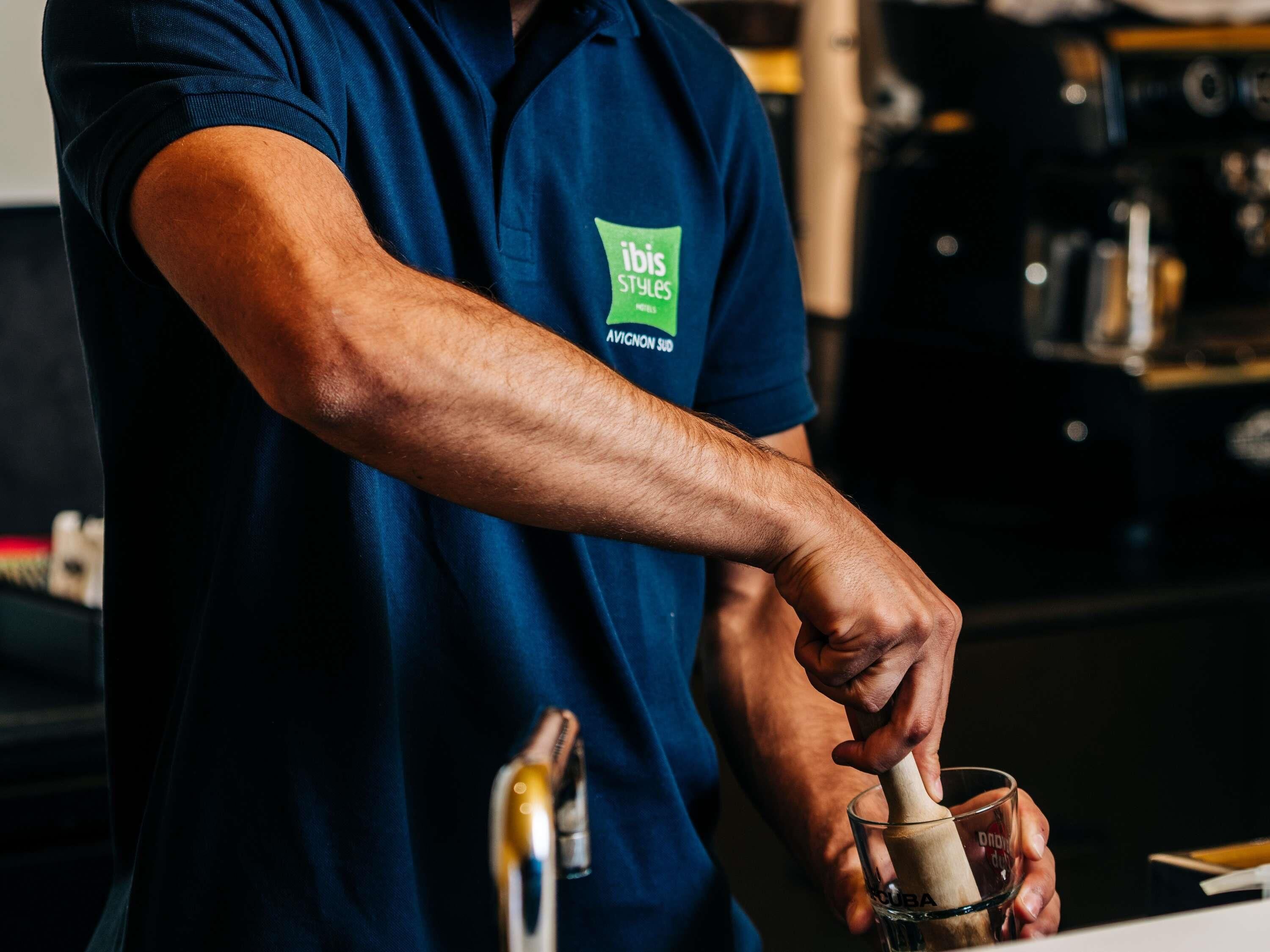A person wearing a blue shirt with a logo, using a wooden tool to mix a drink in a glass.