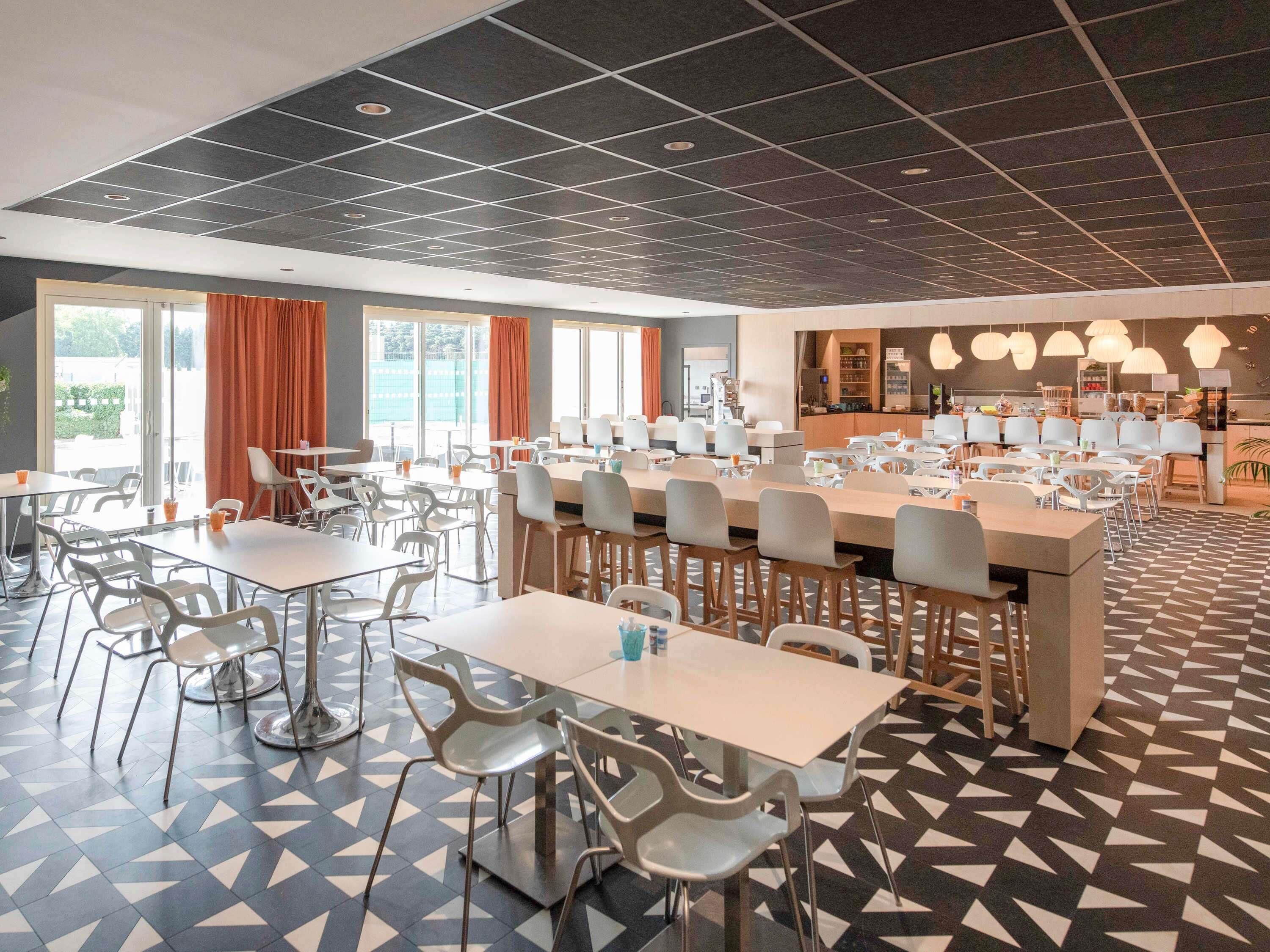 Un espace repas moderne avec plusieurs tables et chaises, un coin cuisine et de grandes fenêtres ornées de rideaux orange.