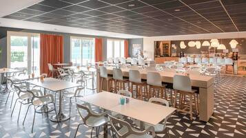 Un espace repas moderne avec plusieurs tables et chaises, un coin cuisine et de grandes fenêtres ornées de rideaux orange.