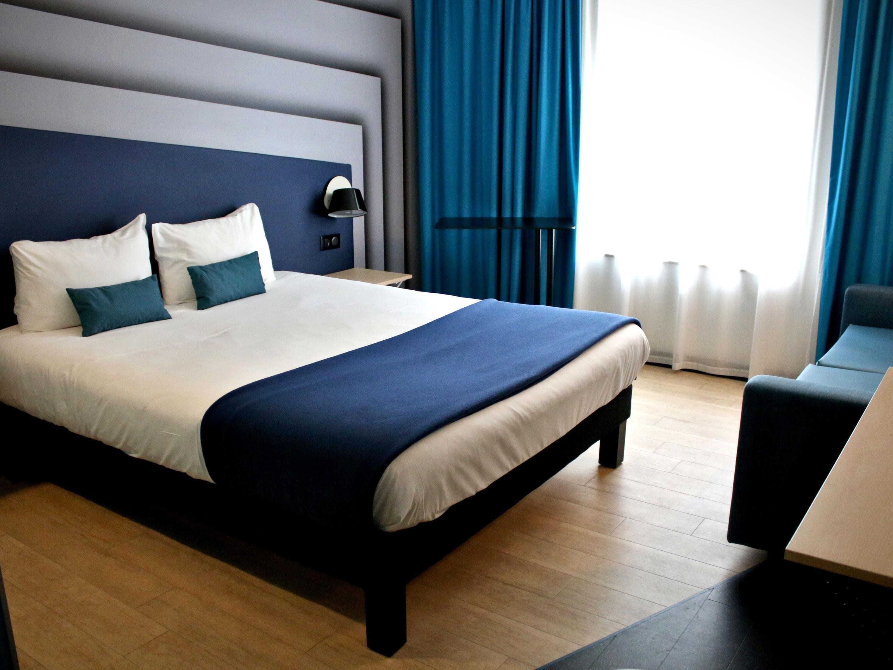 A modern hotel room with a large bed, a bedside table, a lamp, and a view of the outdoors.