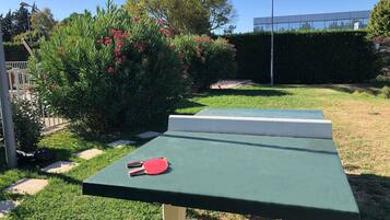 A green outdoor table with a red paddle, surrounded by grass and bushes.