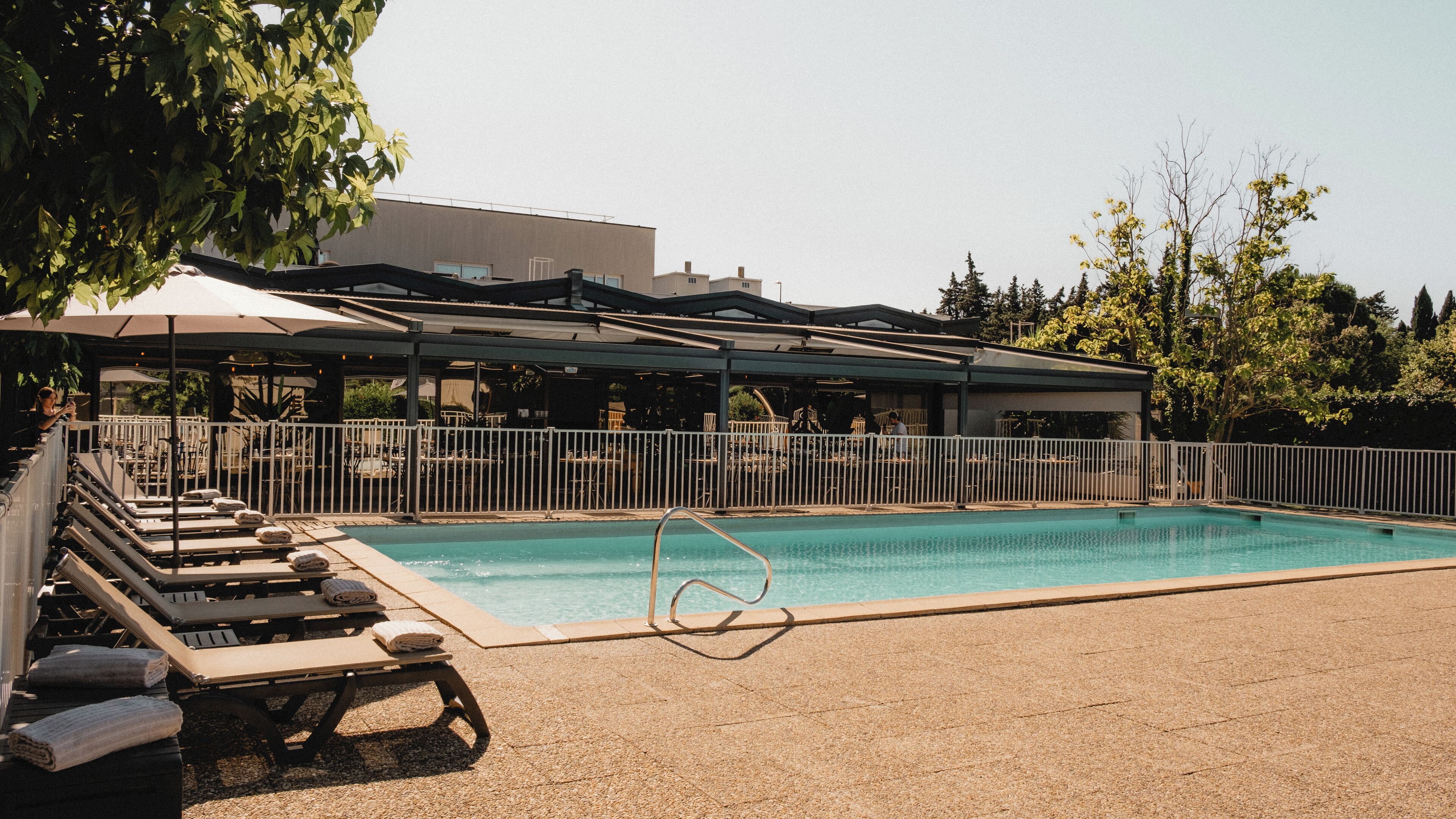 A swimming pool with lounge chairs and umbrellas, surrounded by a fence and trees.