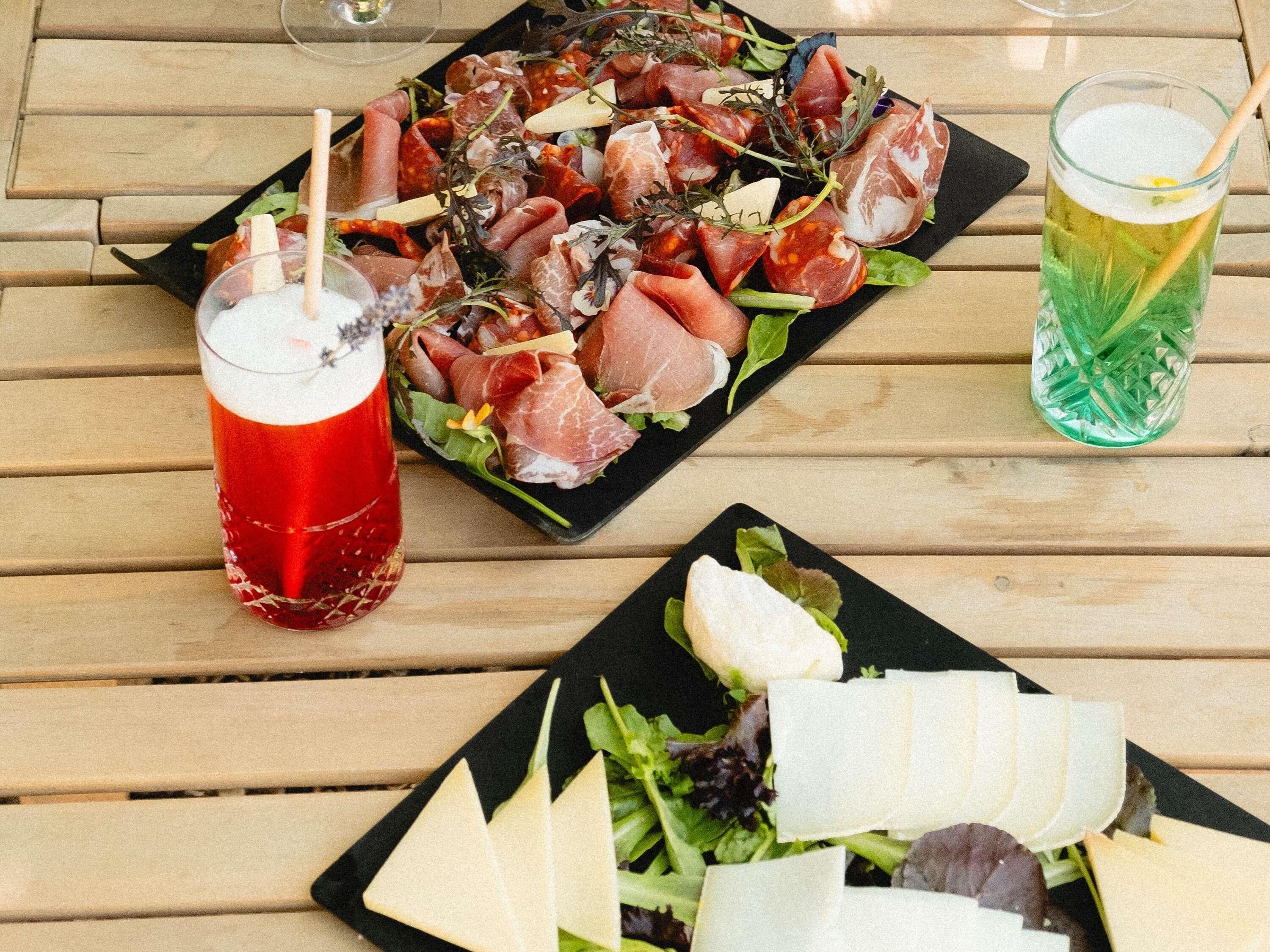 A wooden table with a black plate of assorted meats, a glass of red beverage, and a glass of green beverage.