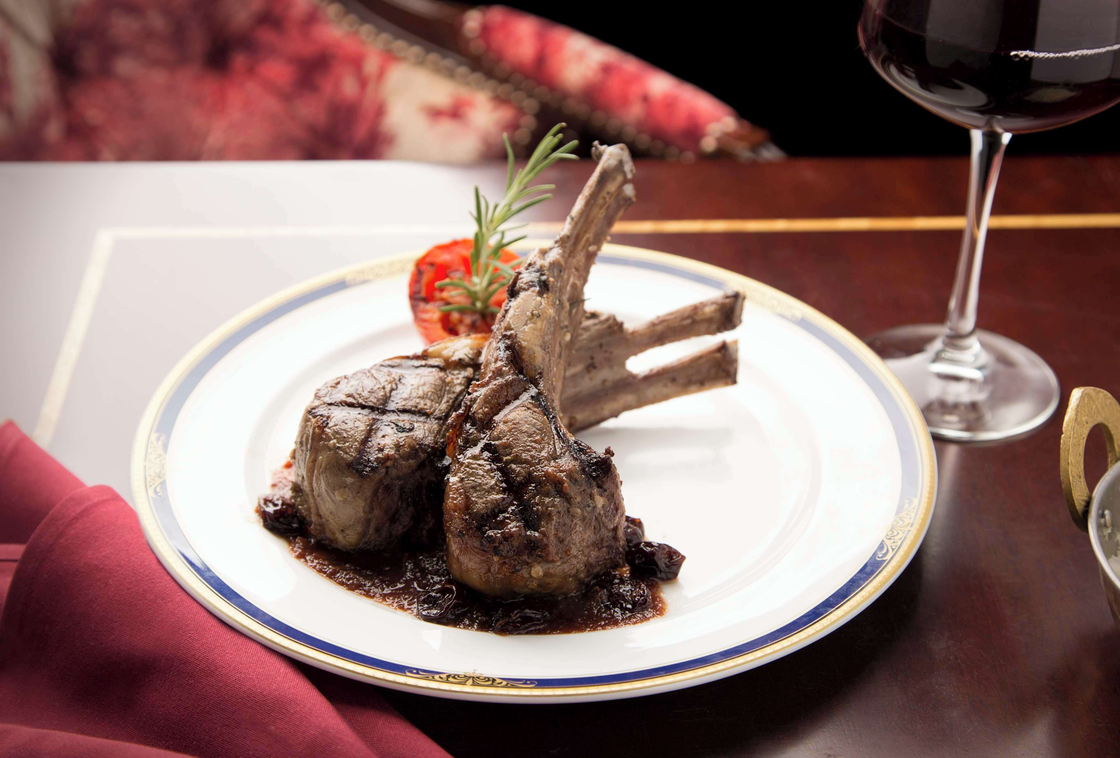 A plate of lamb chops with a garnish, a glass of red wine, and a red napkin on a table.