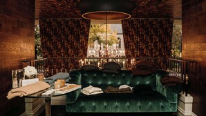 A green velvet sofa with a table beside it, a chandelier, and a view of a building through patterned curtains. - Graduate by Hilton Ann Arbor (Ann Arbor)
