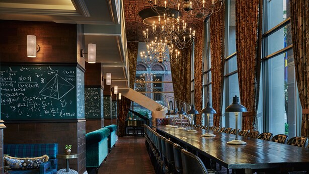 A bar area with a long wooden table, bar stools, a chandelier, and a chalkboard with mathematical equations. - Graduate by Hilton Ann Arbor (Ann Arbor)