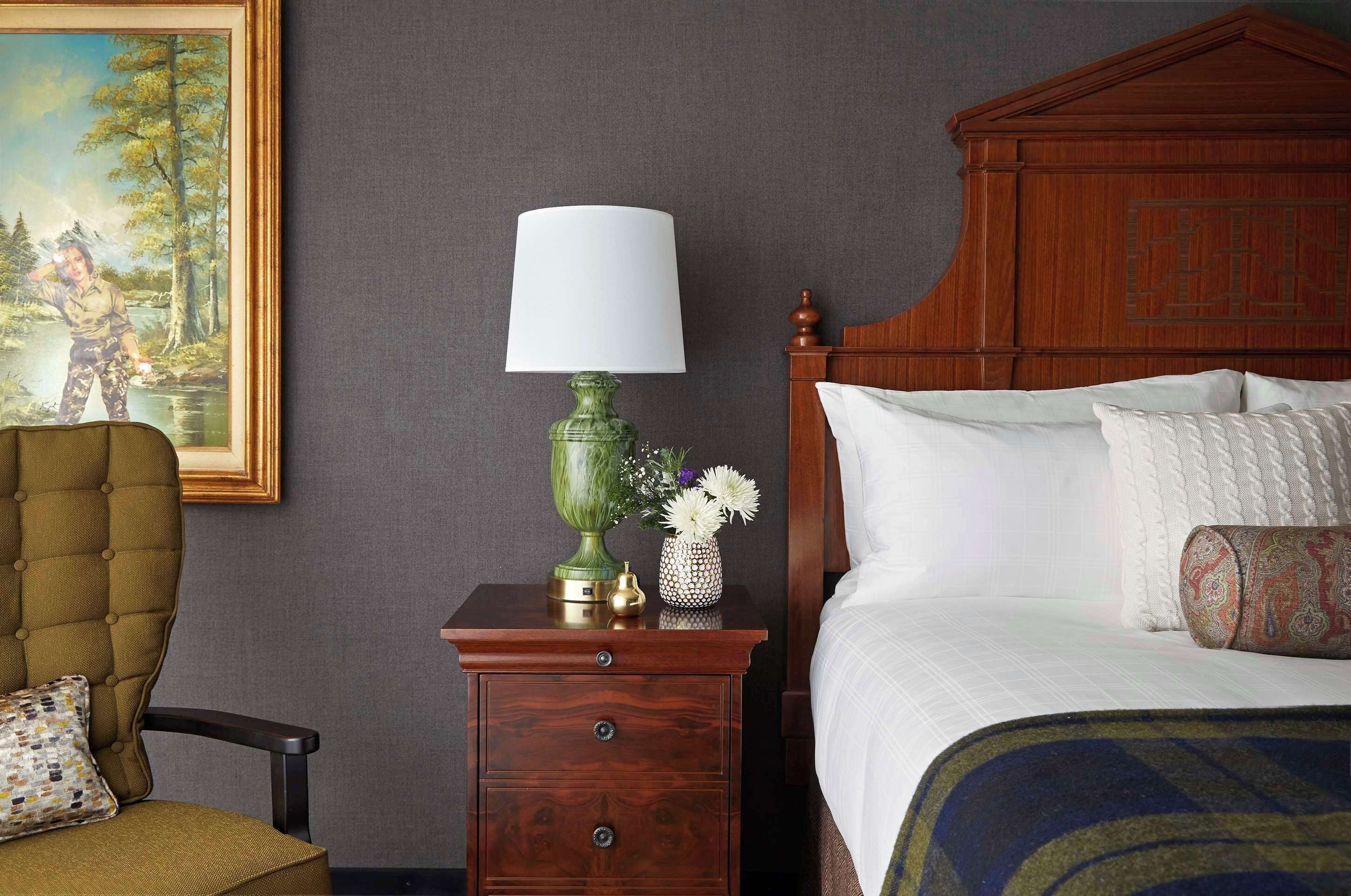 A neatly arranged bedroom with a wooden headboard, a green lamp, a small table with a vase, and a chair with a patterned cushion.