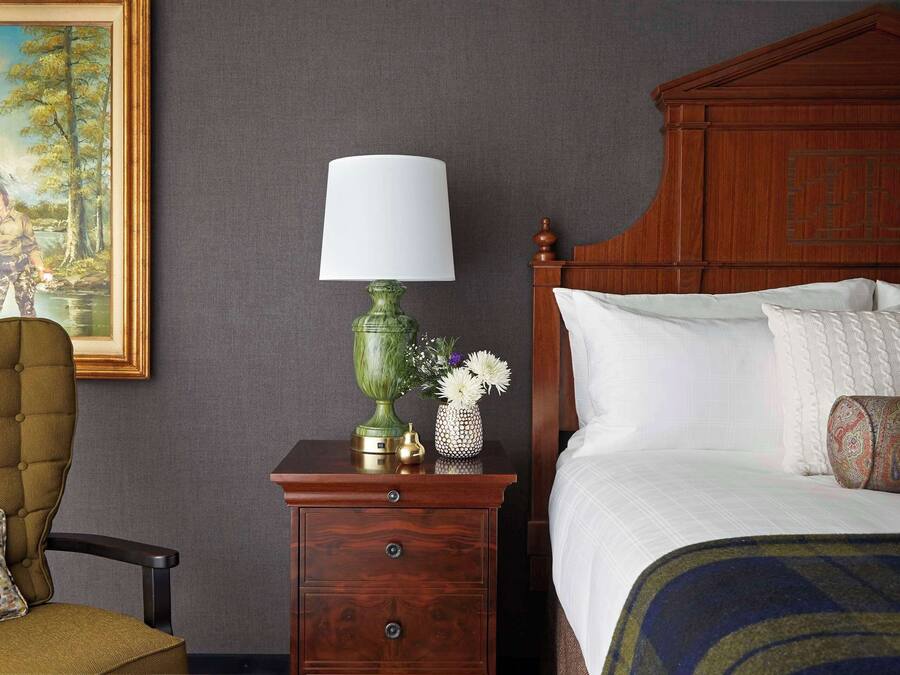 A neatly arranged bedroom with a wooden headboard, a green lamp, a small table with a vase, and a chair with a patterned cushion.