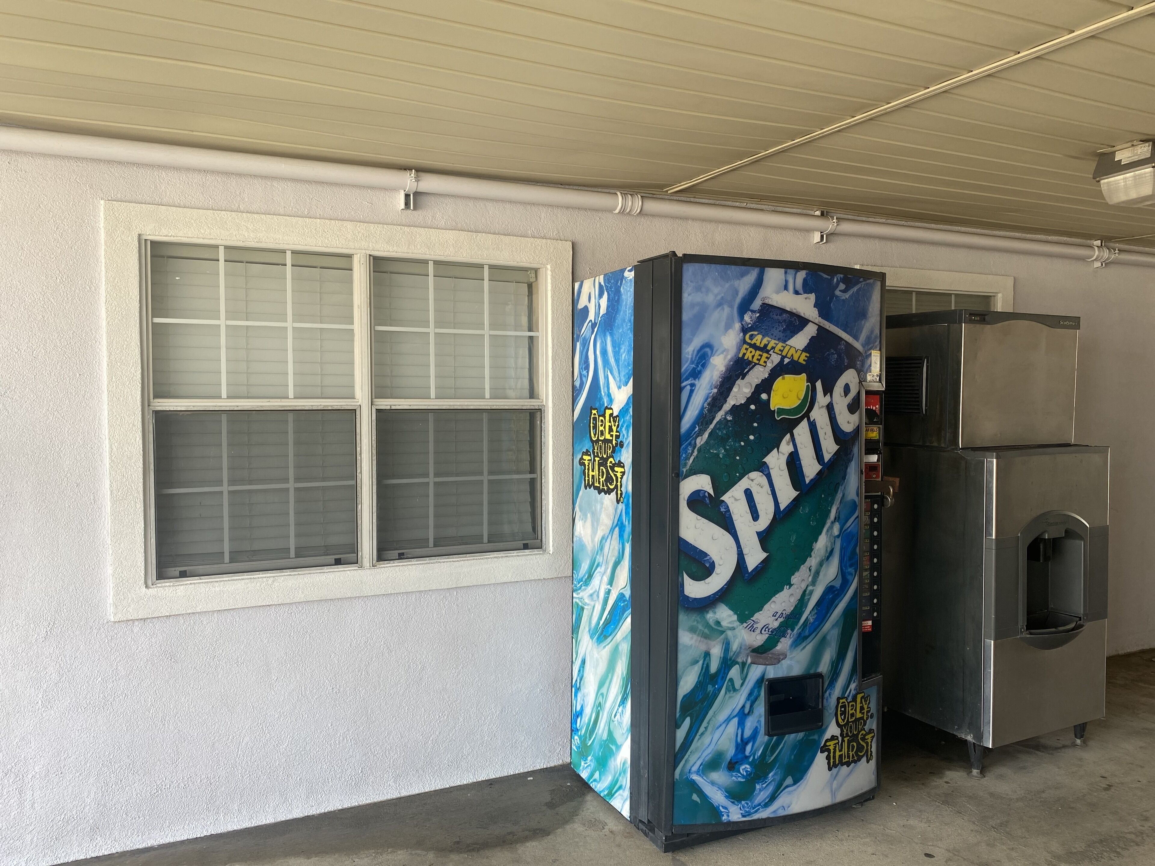 Un distributeur automatique orné du logo Sprite et d’un slogan, placé près d’une fenêtre et d’un appareil en acier inoxydable dans un garage.