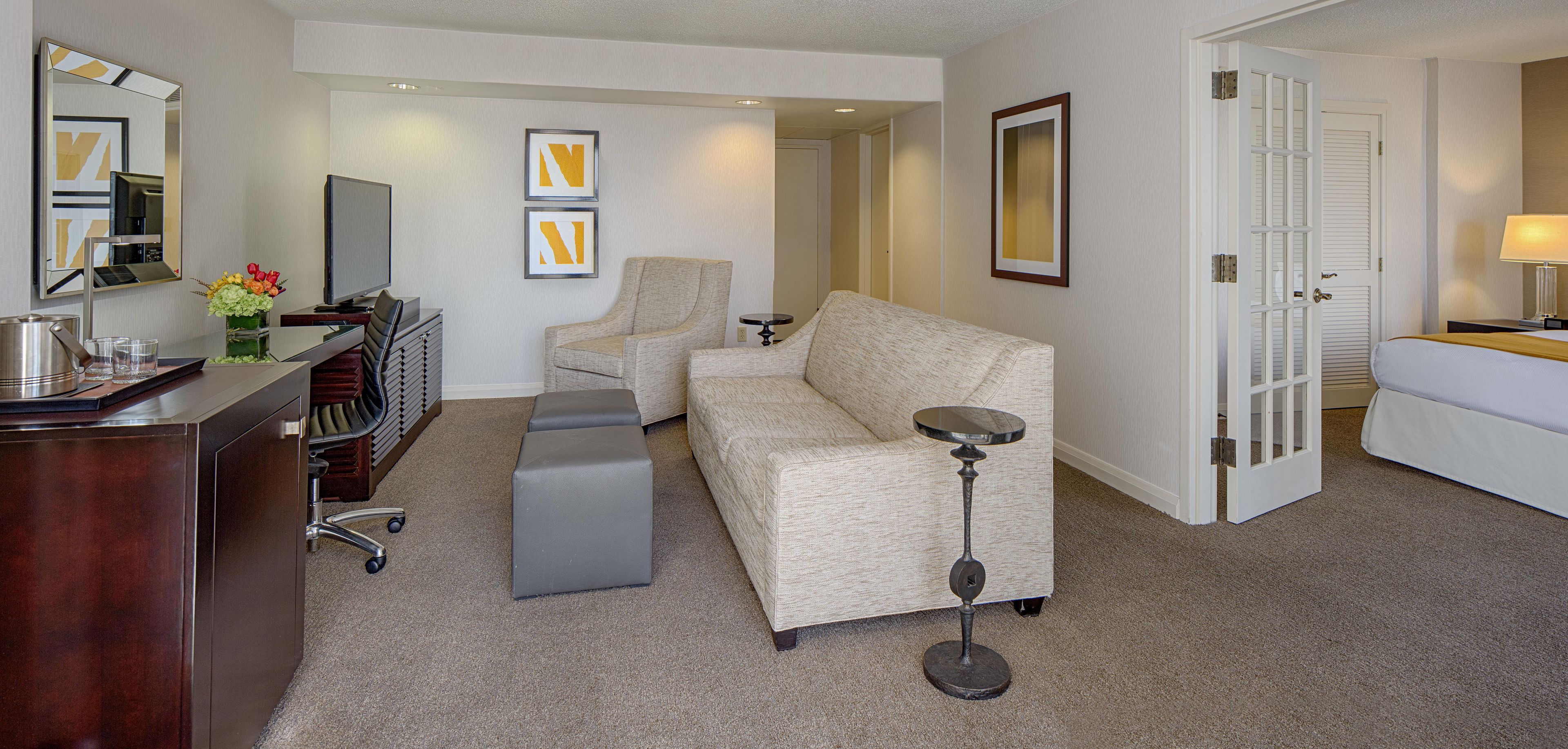 Quarto de hotel com sofá, escrivaninha com computador, cadeira, mesa de cabeceira e luminária.