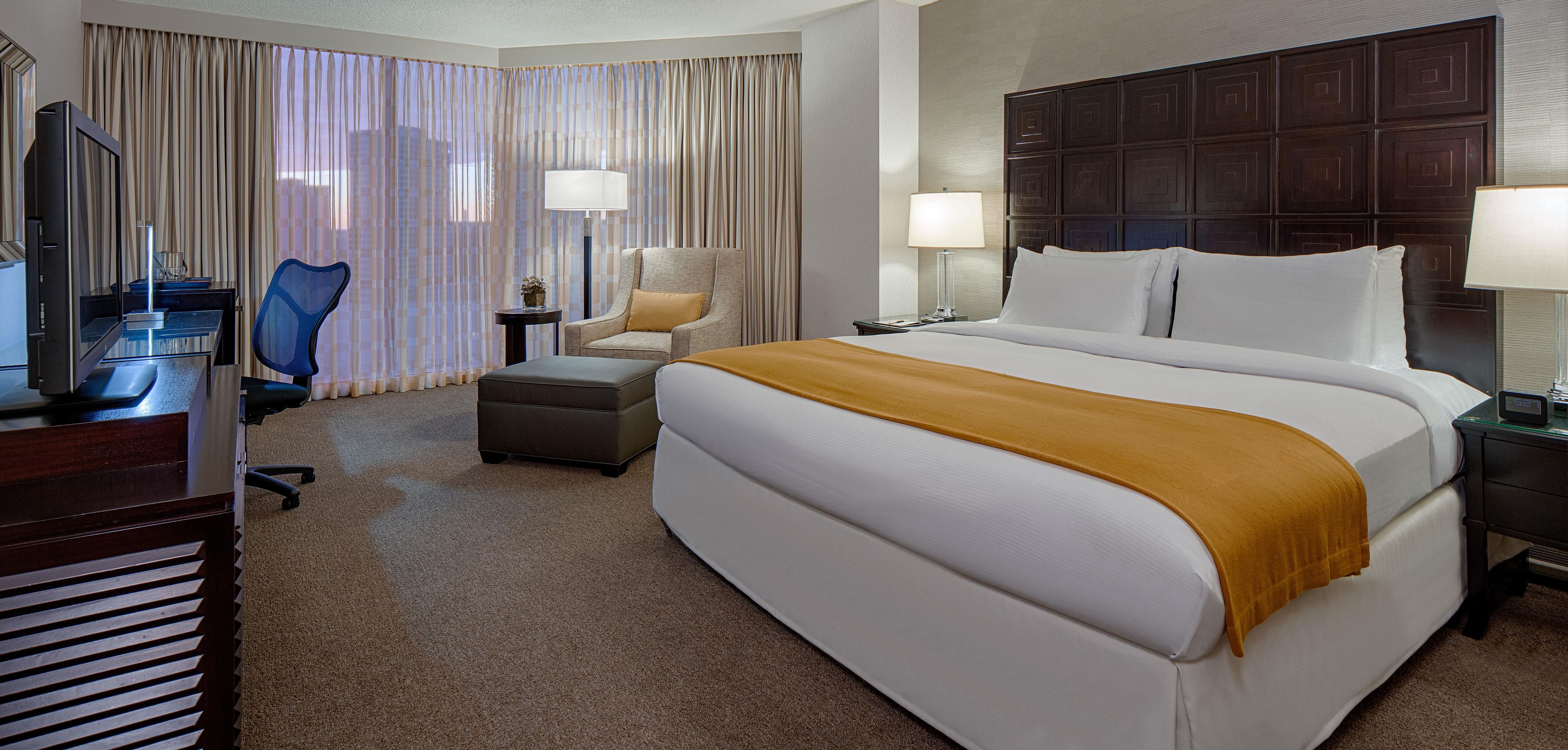 Quarto de hotel com uma cama grande, uma escrivaninha com computador, uma cadeira e uma janela com cortinas.