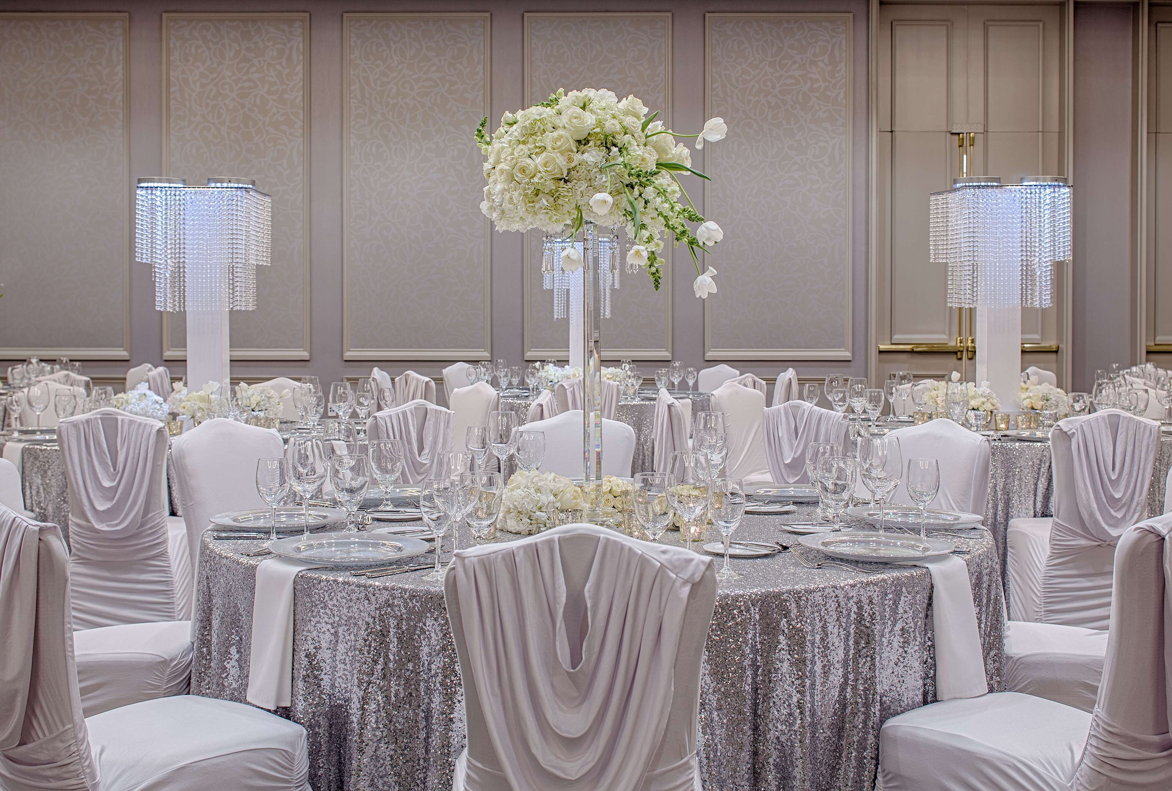 A banquet hall with round tables covered in silver tablecloths, white chairs, and floral centerpieces.