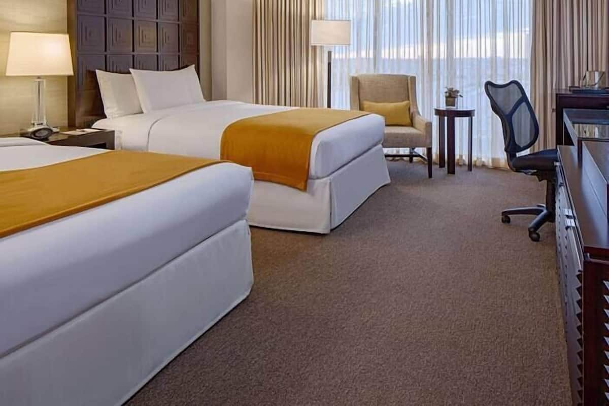 A hotel room with two beds, a desk, a chair, and a view of the outdoors.