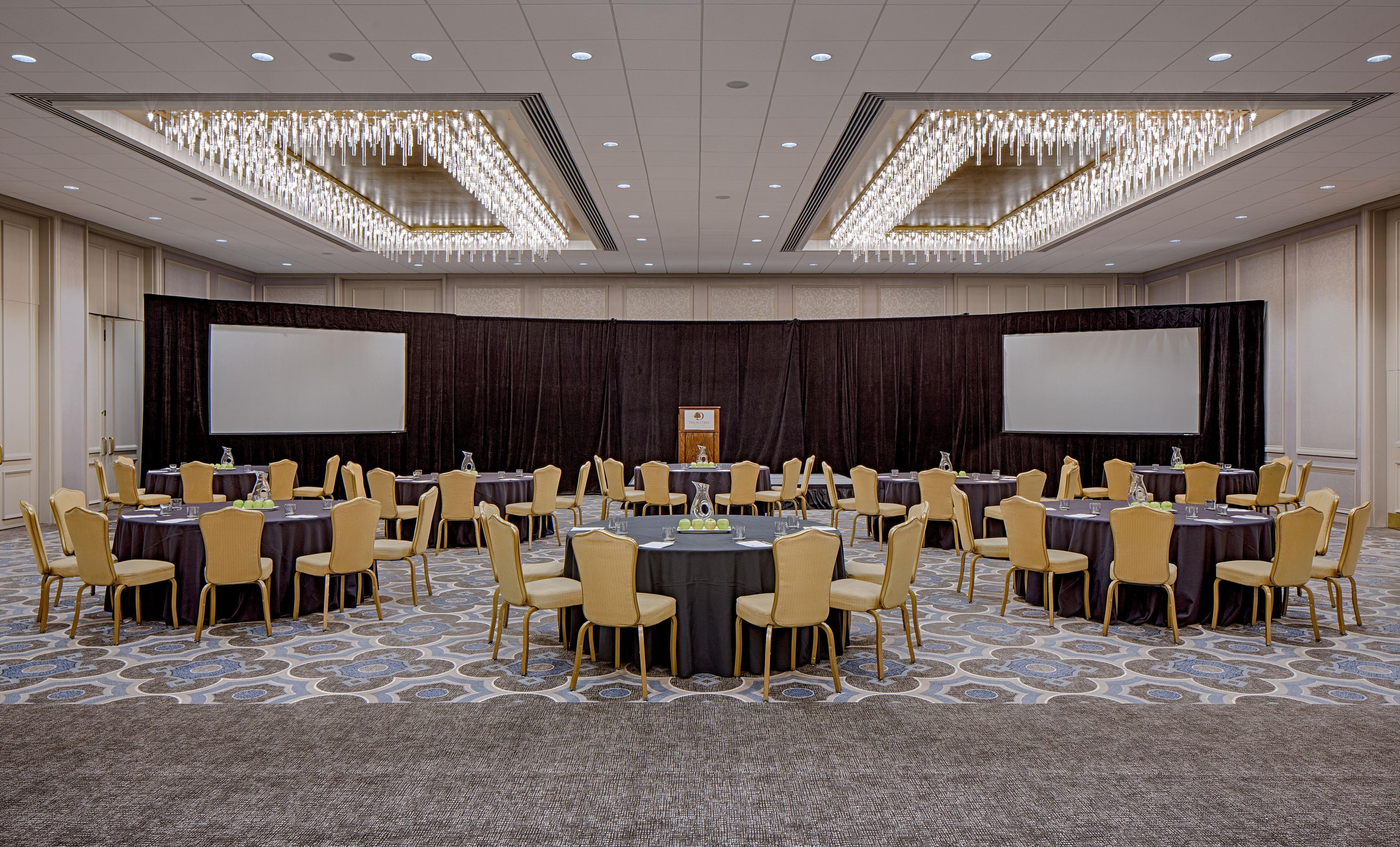 A conference room with round tables, chairs, a stage with a screen, and a chandelier.