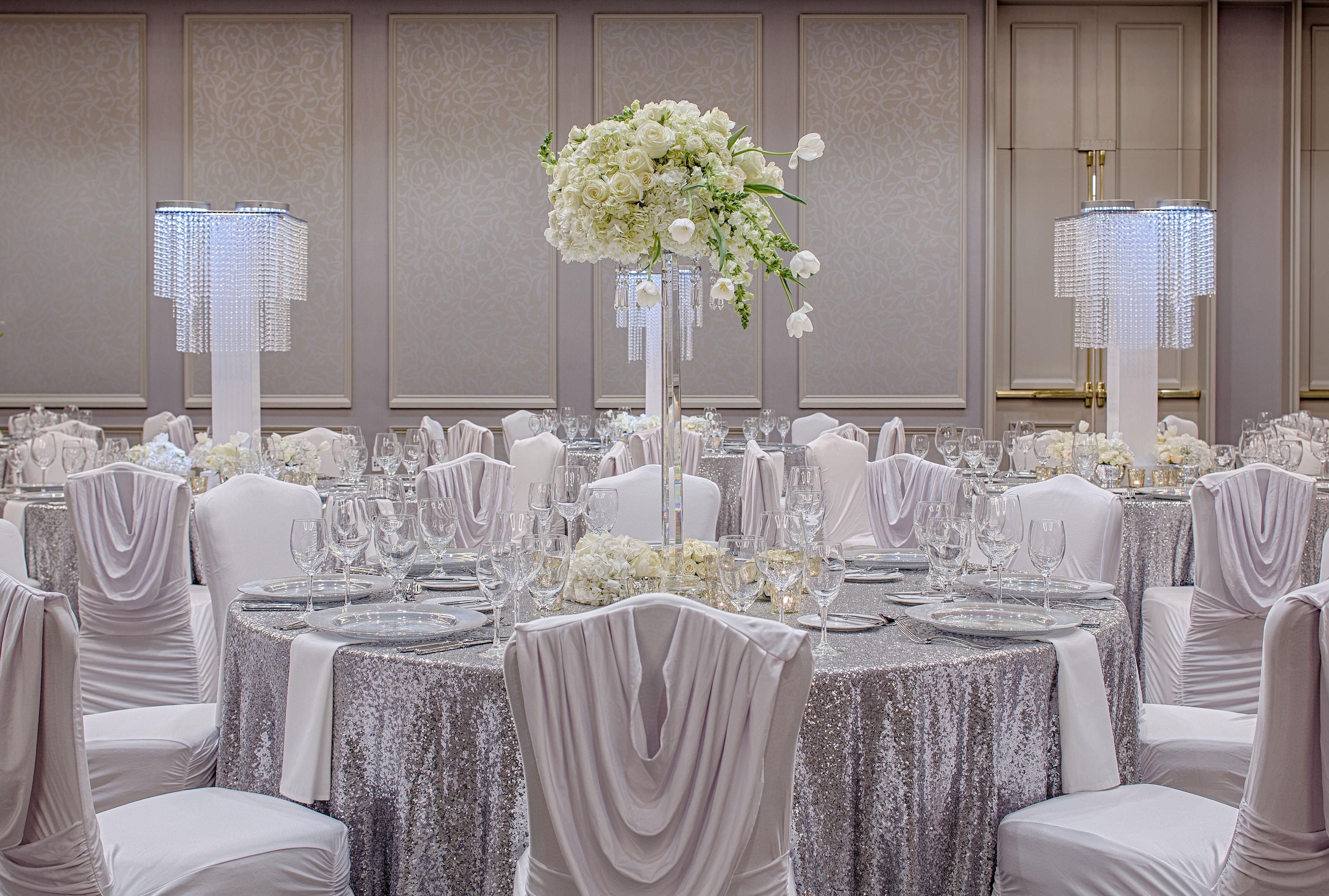 Une salle de banquet avec des tables rondes dressées pour un événement formel, agrémentée de lustres en cristal et de compositions florales blanches.