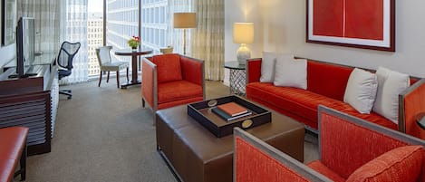 Een hotelkamer met een rode bank, een bruin pouf, een bureau met een computer, een stoel en uitzicht op de stad door grote ramen.