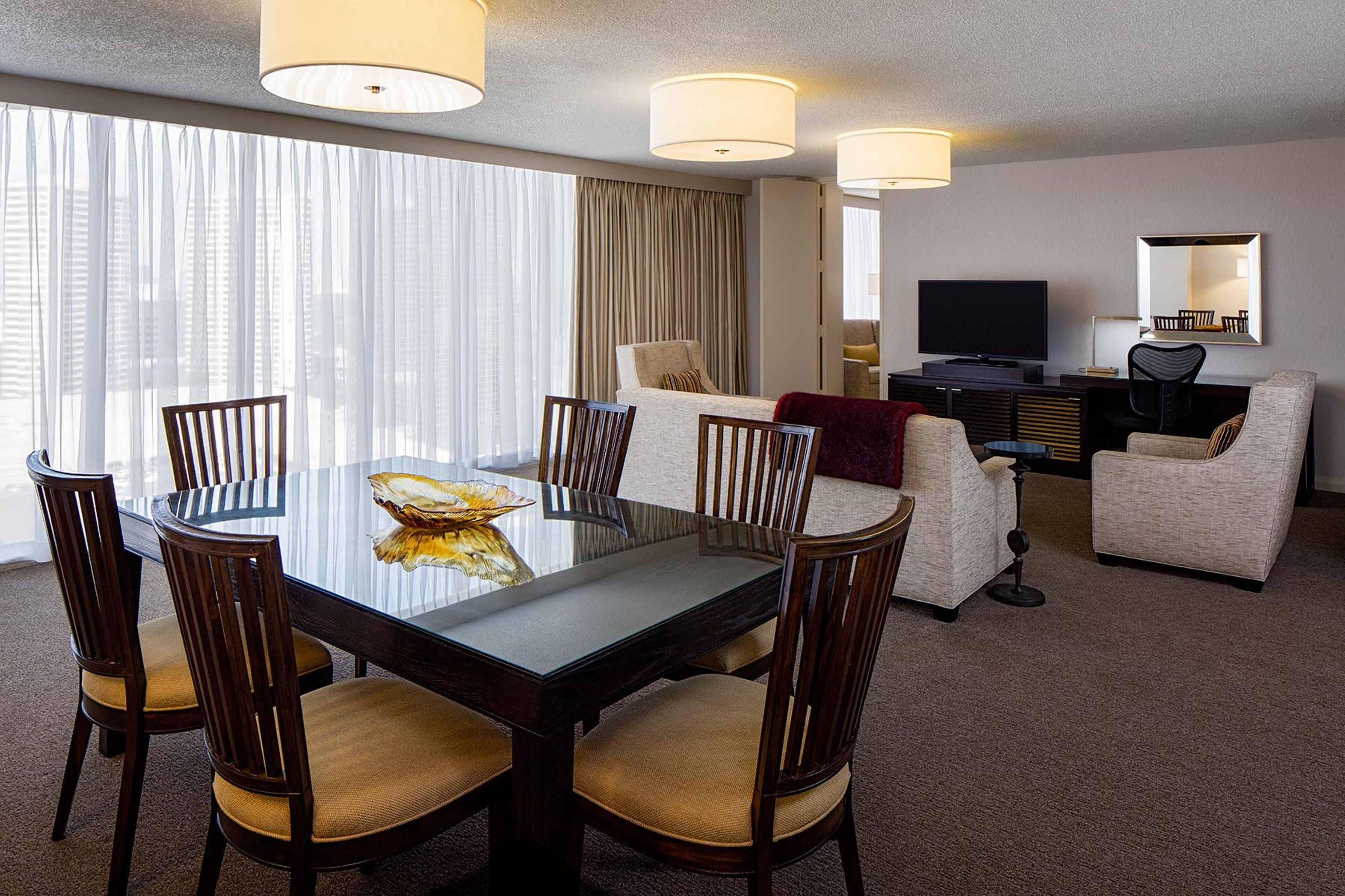 A hotel room with a dining table, chairs, a sofa, a TV, and a large window with curtains.