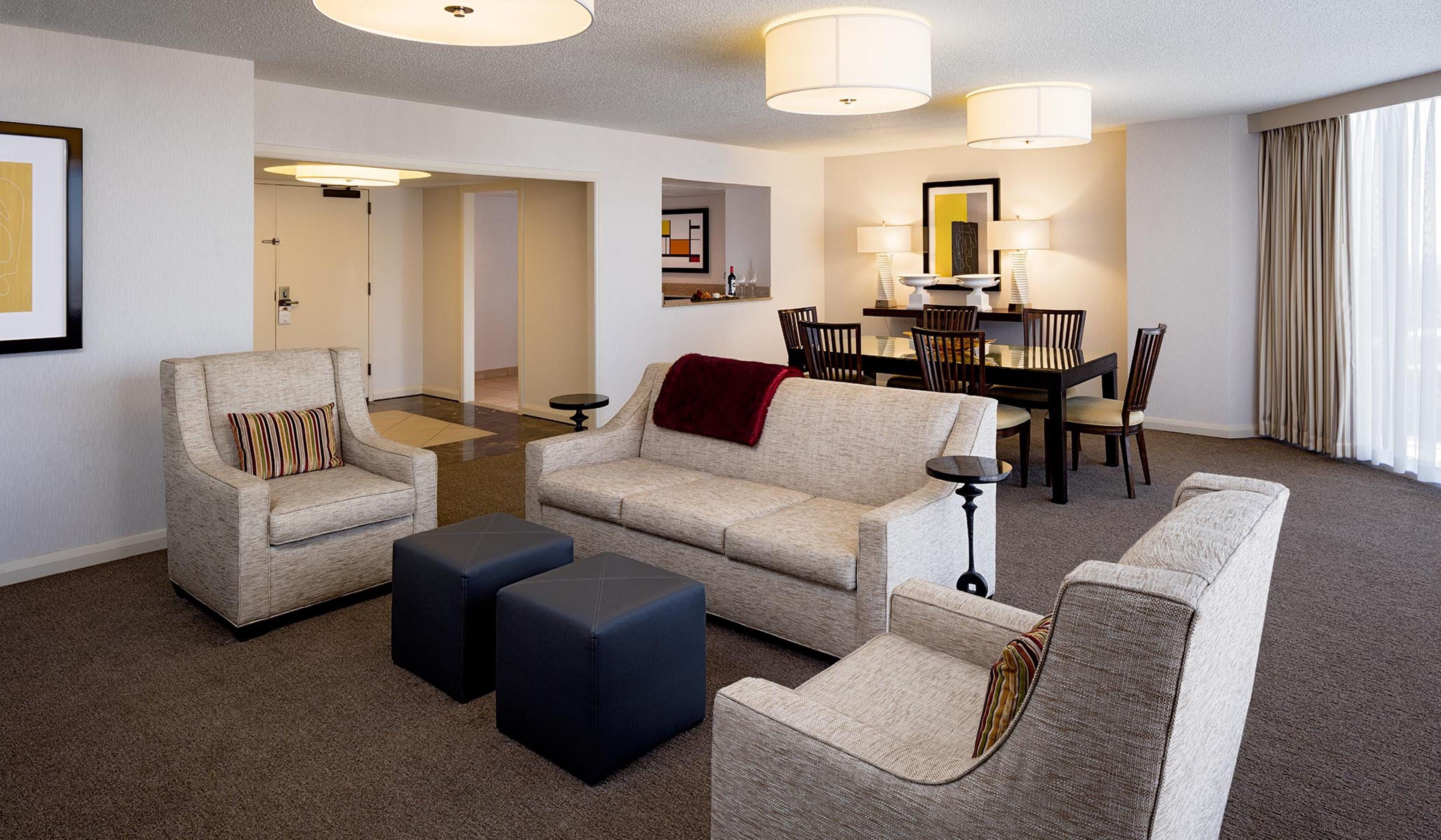 A hotel room with a living area, a dining table with chairs, a sofa, two ottomans, and a small side table.