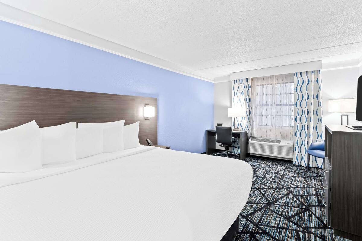 Une chambre d’hôtel avec un grand lit, un bureau, une chaise, une lampe de bureau, une fenêtre avec des rideaux et un climatiseur fixé au mur.