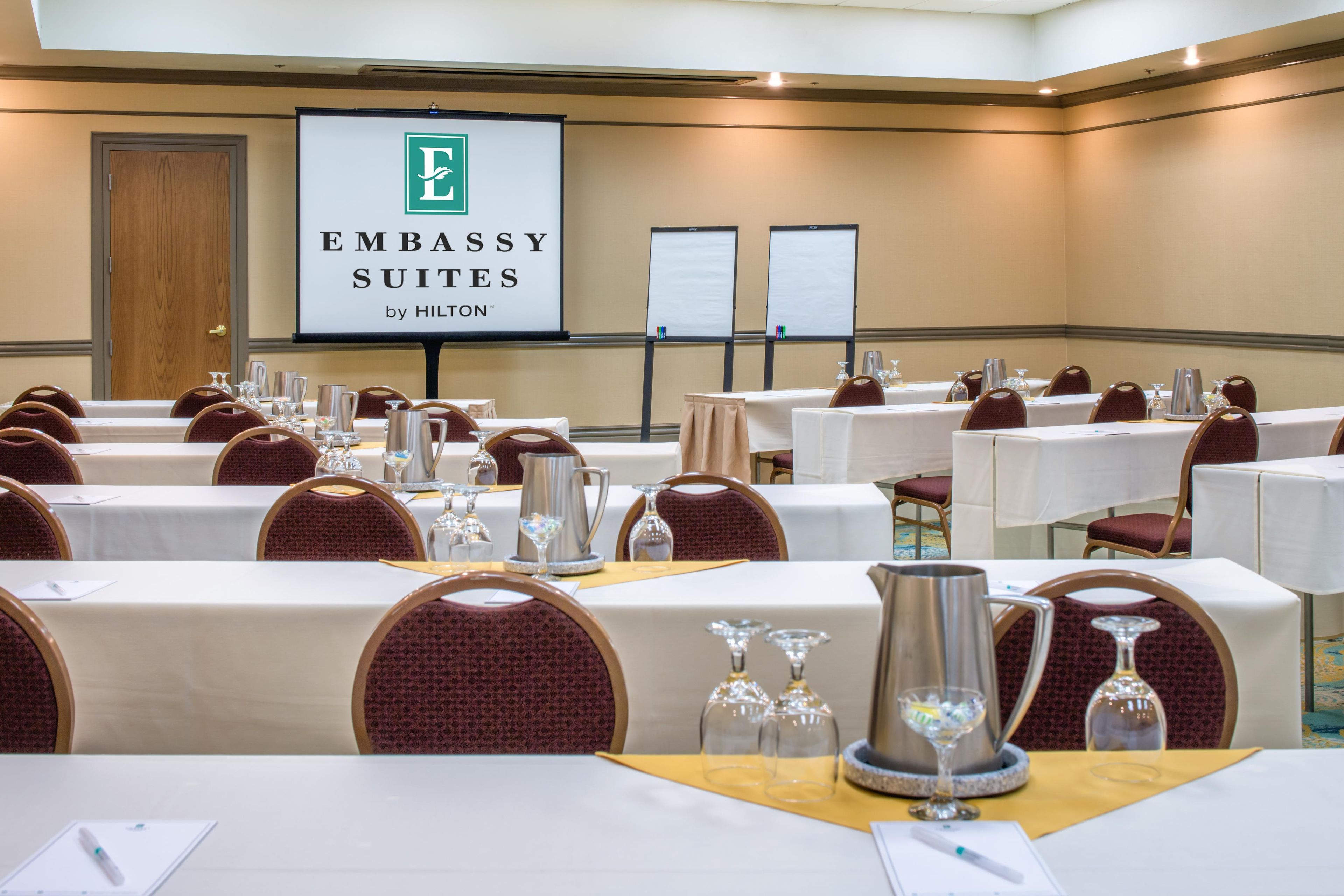 A conference room with tables set for a meeting, a projector screen displaying 'Embassy Suites by Hilton', and a door to the left.