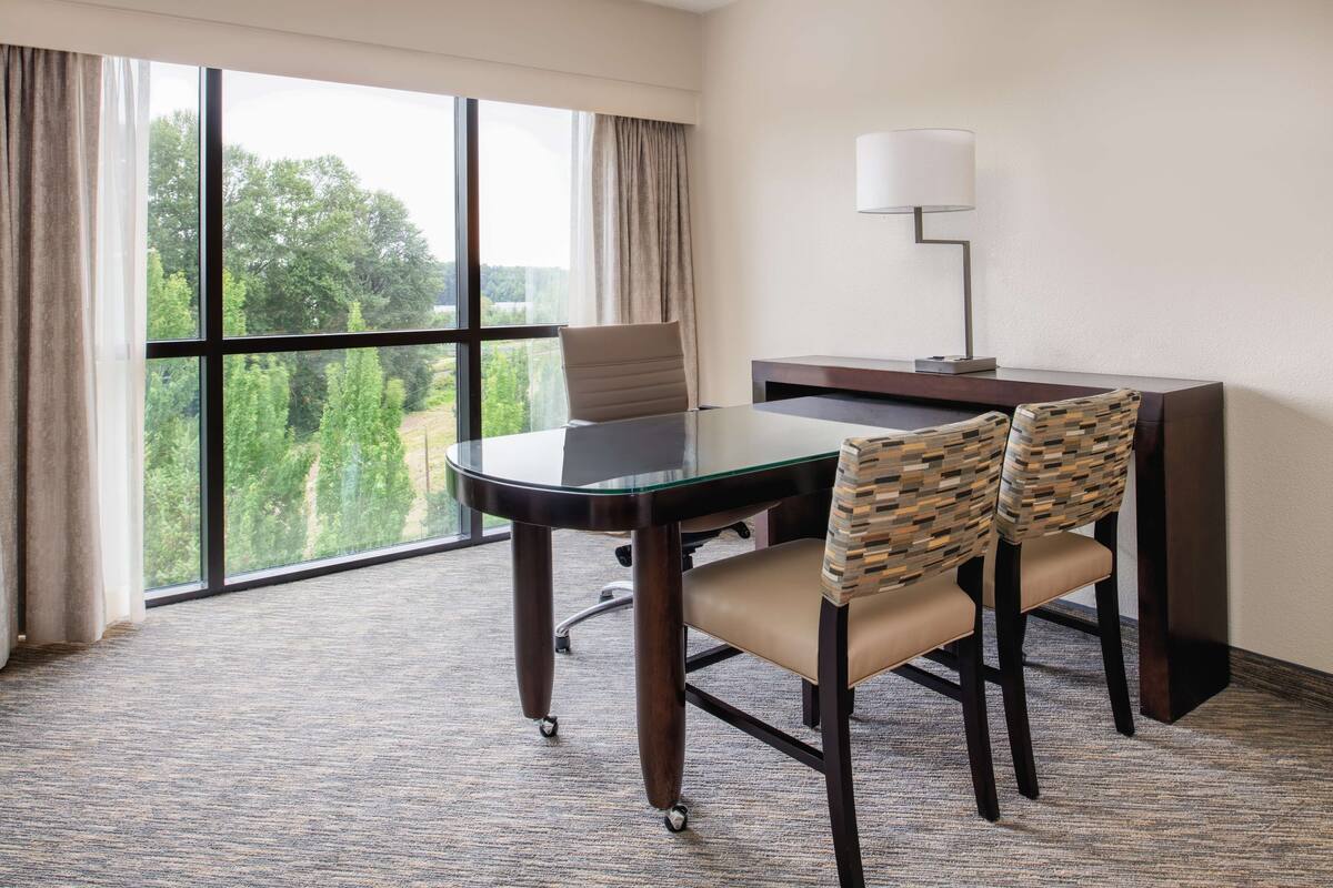 A hotel room with a large window, a glass table, two chairs, a desk, and a lamp.