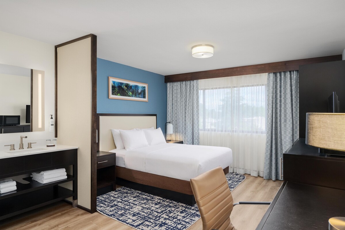 A hotel room with a bed, a desk with a computer, a television, a bathroom with a mirror, and a window with curtains.