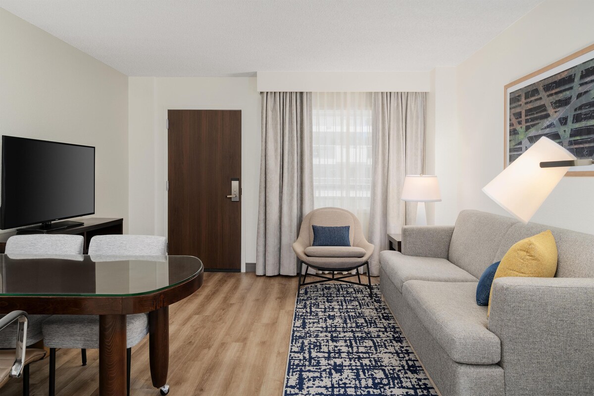 A hotel room with a flat-screen TV, a dining table with chairs, a sofa, a patterned rug, and a window with curtains.
