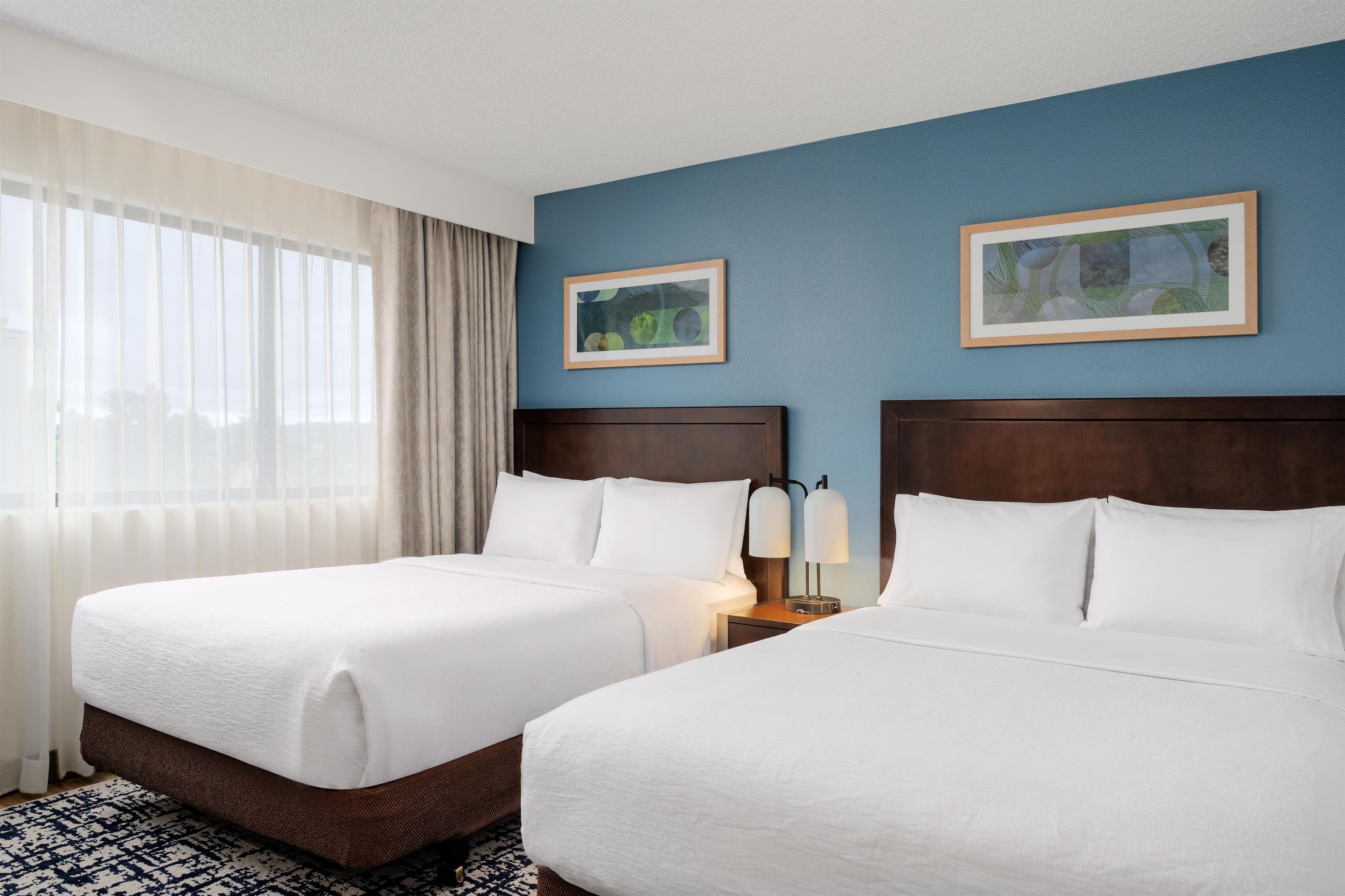 Quarto de hotel com duas camas, uma mesa de cabeceira com abajur, uma janela com cortinas e duas quadros na parede.