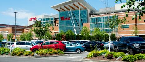 A modern building with a large glass facade, a parking lot with various cars, and a landscaped area with shrubs and trees.