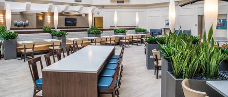 A spacious indoor area with multiple seating arrangements, including a long communal table and several smaller tables with chairs. There are potted plants placed around the seating areas.