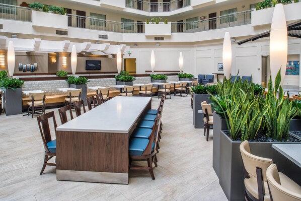 A spacious indoor area with multiple seating arrangements, including a long communal table and several smaller tables with chairs. There are potted plants placed around the seating areas.