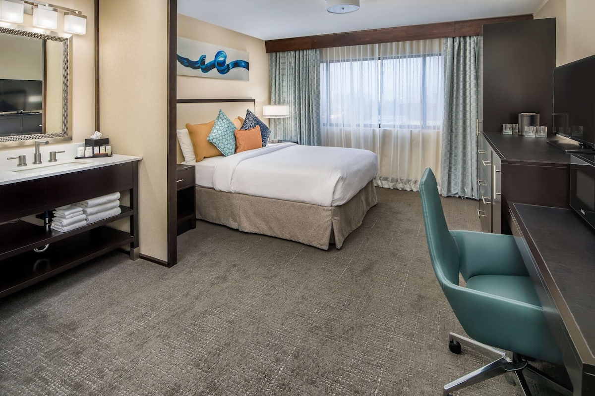 A hotel room with a bed, a desk, a chair, a vanity with a mirror, and a television.