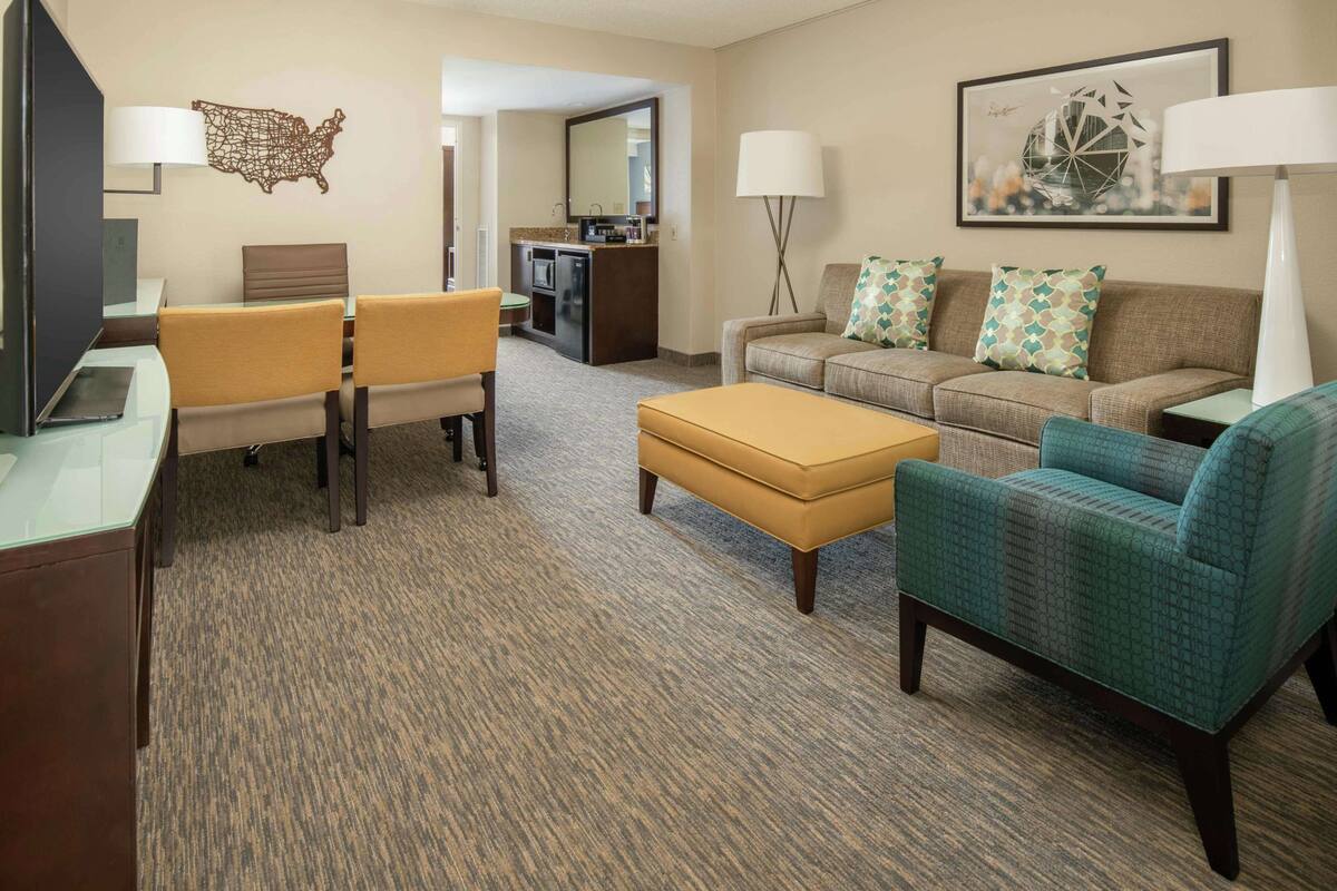 A hotel room with a dining area, a sofa set, a coffee table, a TV, a desk, and a lamp.