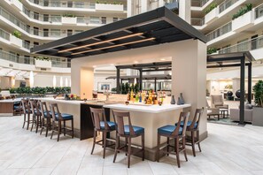 Un bar moderno con una barra central, diversas opciones de asientos y vistas al interior del edificio circundante.