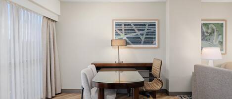 Habitación de hotel con escritorio de madera, una silla, un sofá, una lámpara y un ventanal con cortinas.