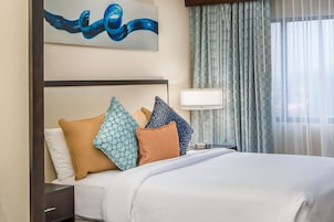 Una habitación de hotel con una cama bien hecha, una mesita de noche con lámpara y un ventanal con cortinas.