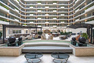 Un vestíbulo de hotel de varios niveles con piscina central, zonas de estar y balcones con plantas.