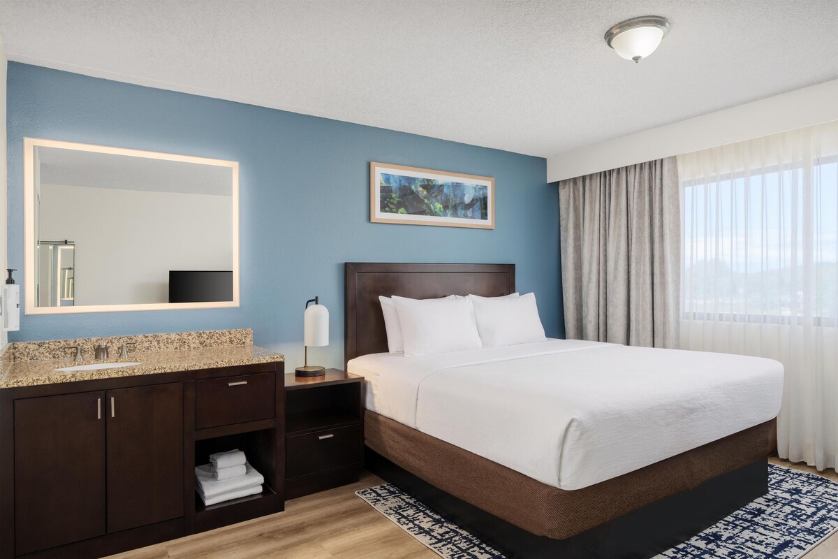 A hotel room with a bed, a nightstand, a mirror, a sink, and a window with curtains.