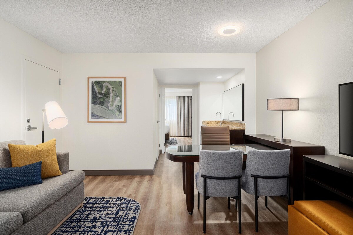 A modern hotel room with a sofa, a dining table, two chairs, a TV, and a bathroom visible through an open door.
