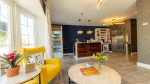Un salon moderne avec un canapé jaune, une table basse ronde sur laquelle sont posés un livre et des fleurs, un coin cuisine équipé d’un réfrigérateur et d’un four à micro-ondes, et une grande fenêtre donnant sur l’extérieur.