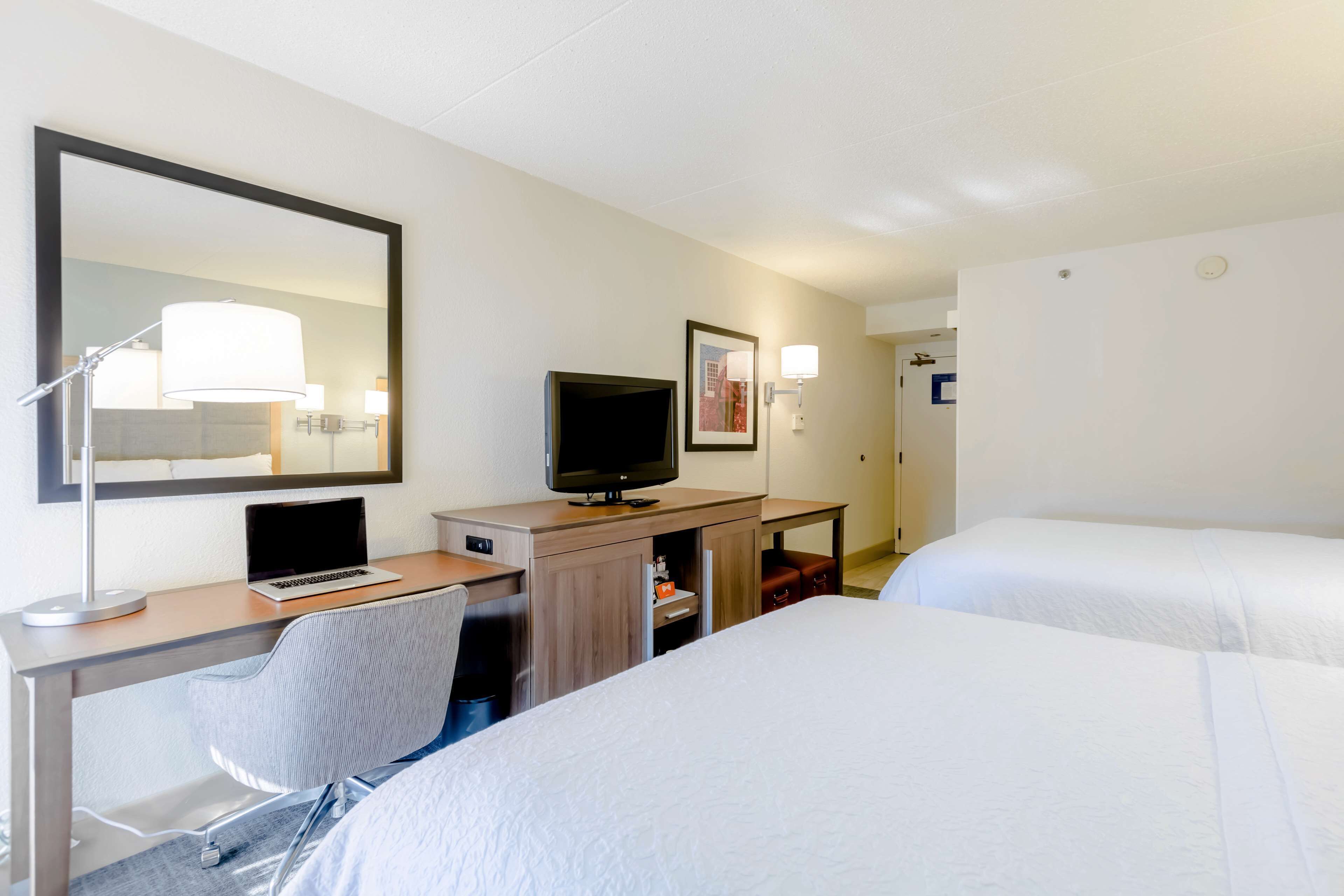 A hotel room with a desk, a television, a chair, and two beds.