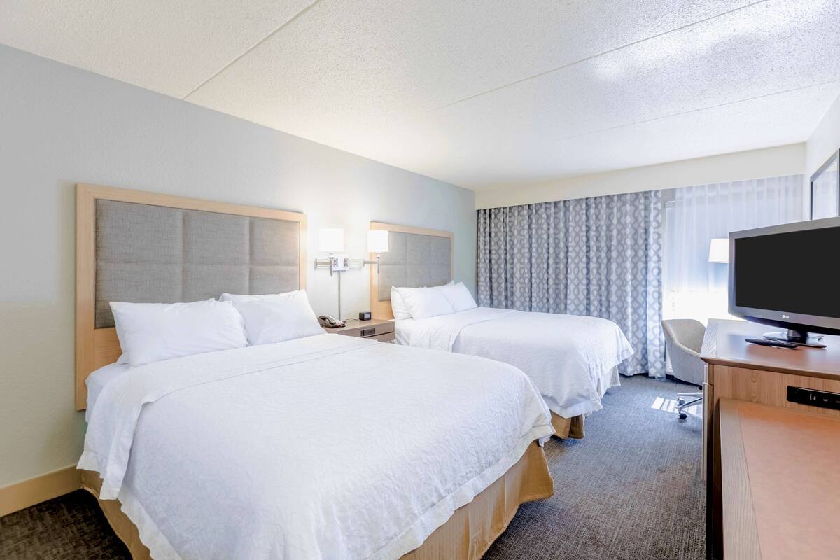 A hotel room with two beds, a desk, a TV, and a window with curtains.