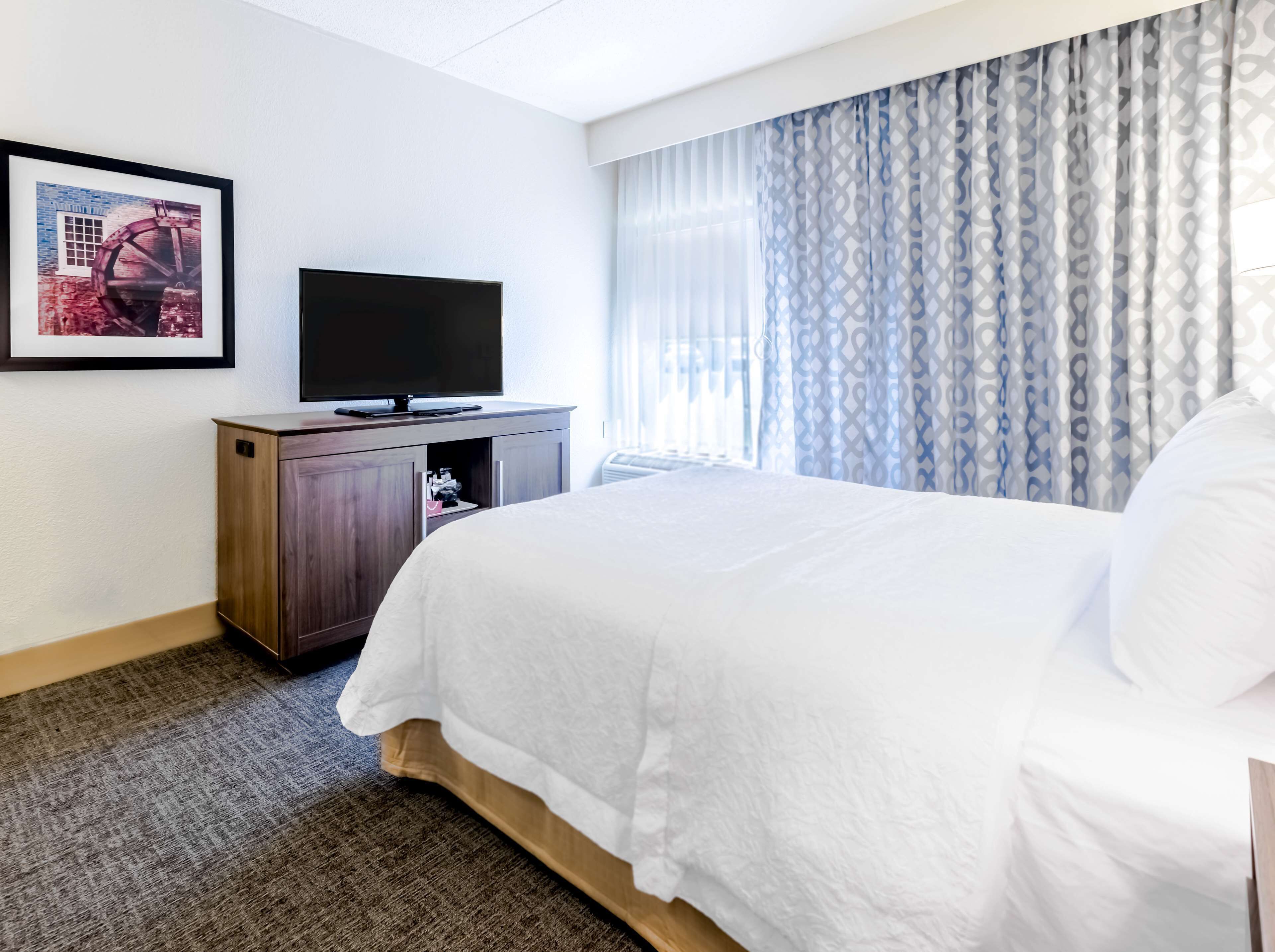 Une chambre d’hôtel avec un lit, un téléviseur posé sur une commode en bois et un tableau encadré accroché au mur.