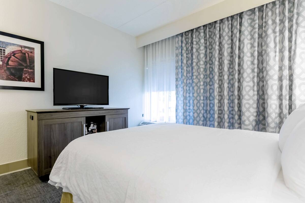 A hotel room with a bed, a television on a wooden cabinet, and a window with patterned curtains.