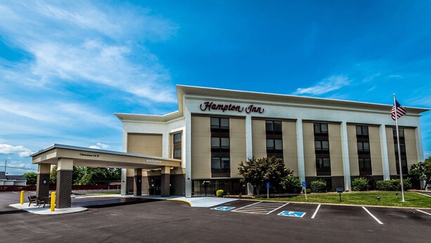 A Hampton Inn hotel with a clear sky, a parking lot with handicapped accessible spaces, and an American flag.