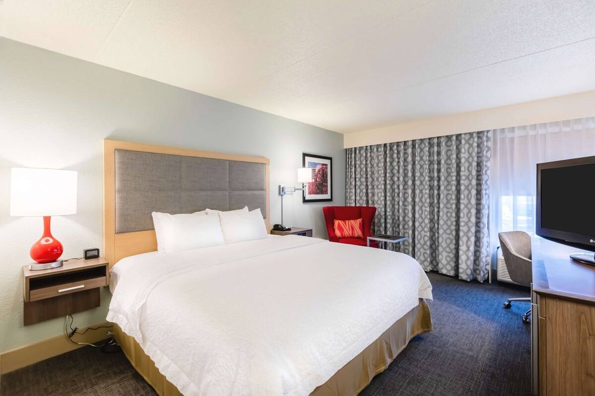 A hotel room with a large bed, a desk with a television, a red chair, and a window with curtains.